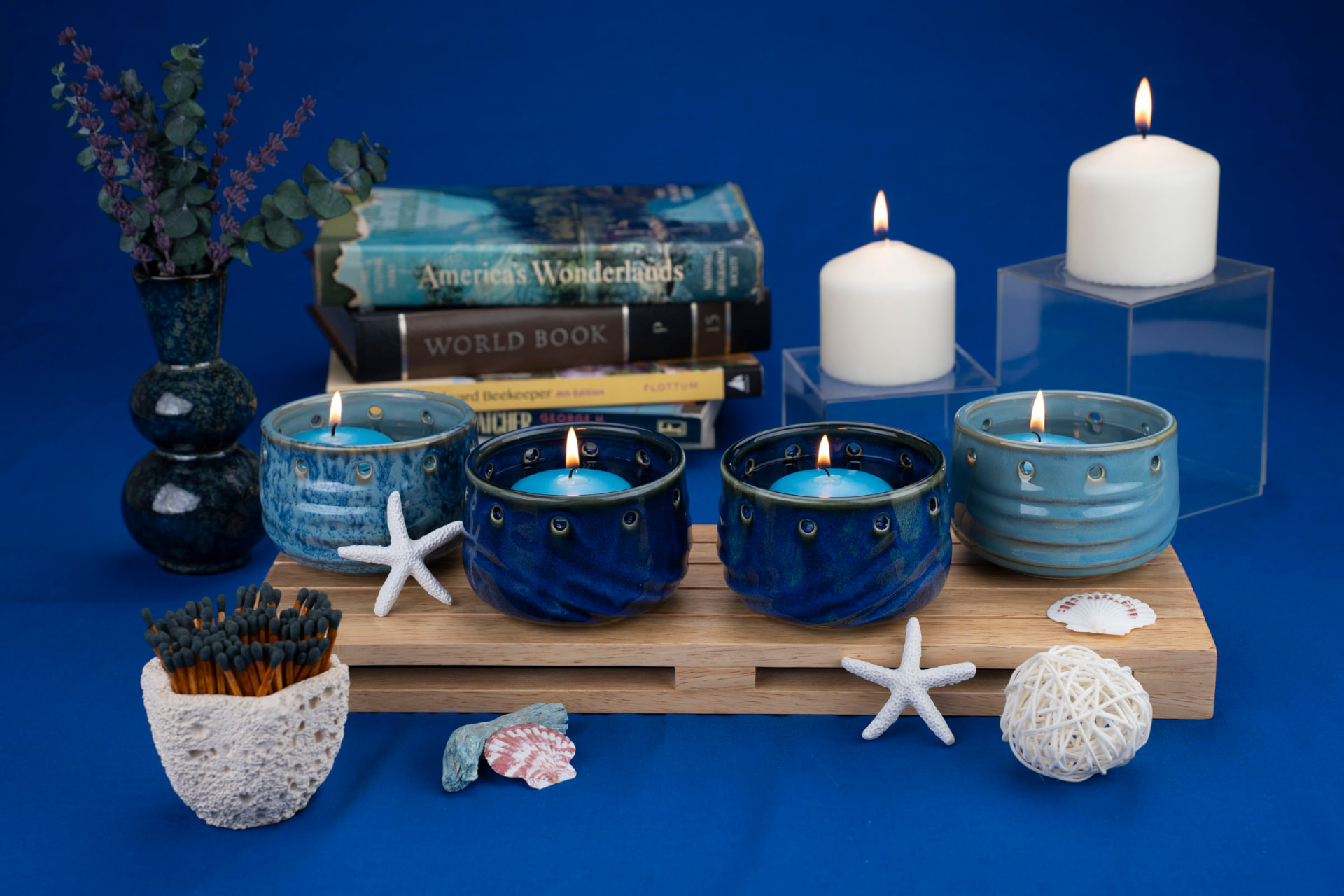 Four lit ceramic candle vessels in blue and teal glazes arranged on a wooden tray with seashells and books against a blue backdrop