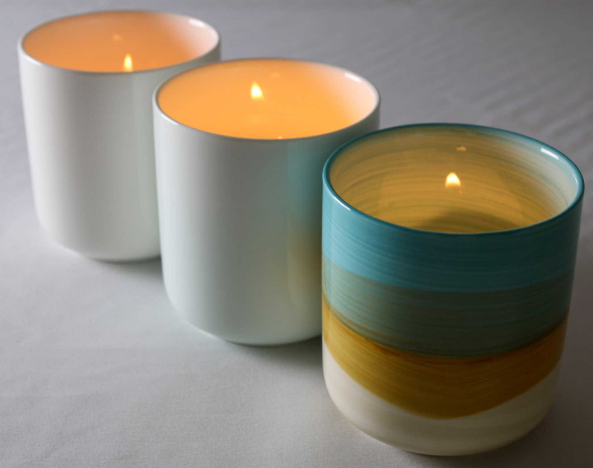 Three lit ceramic candle vessels in cream, white, and blue-striped glazes