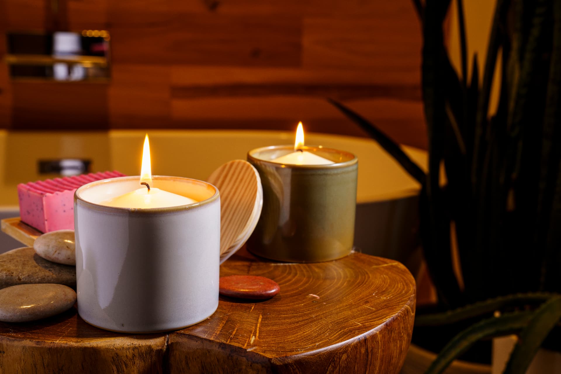 Two lit ceramic candle vessels on a wooden stump with soap and stones in a warm spa setting